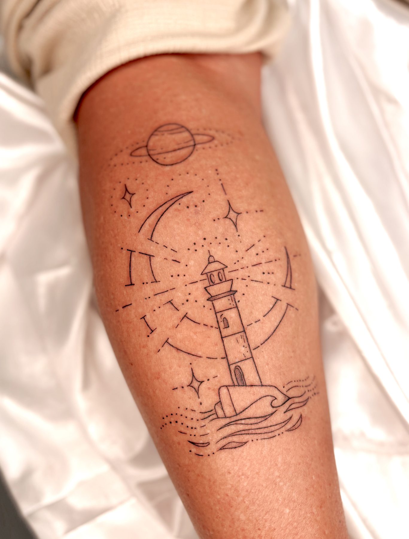 Tatuagem fineline de farol e elementos celestes no antebraço